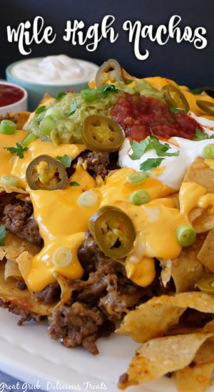 Mile High Nachos are a delicious recipe that is loaded with traditional nacho toppings, but baked in the oven in a deep bowl, creating a delicious nacho mound of goodness. A close up of nachos on a white plate.