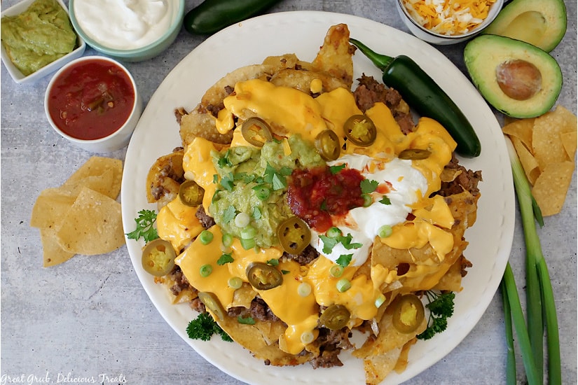 Mile High Nachos are the perfect game day recipe that everyone will enjoy. An over view picture of guacamole, sour cream, salsa, shredded cheese in small white bowls, with avocado, jalapenos, green onions and tortilla chips surrounding a white plate.