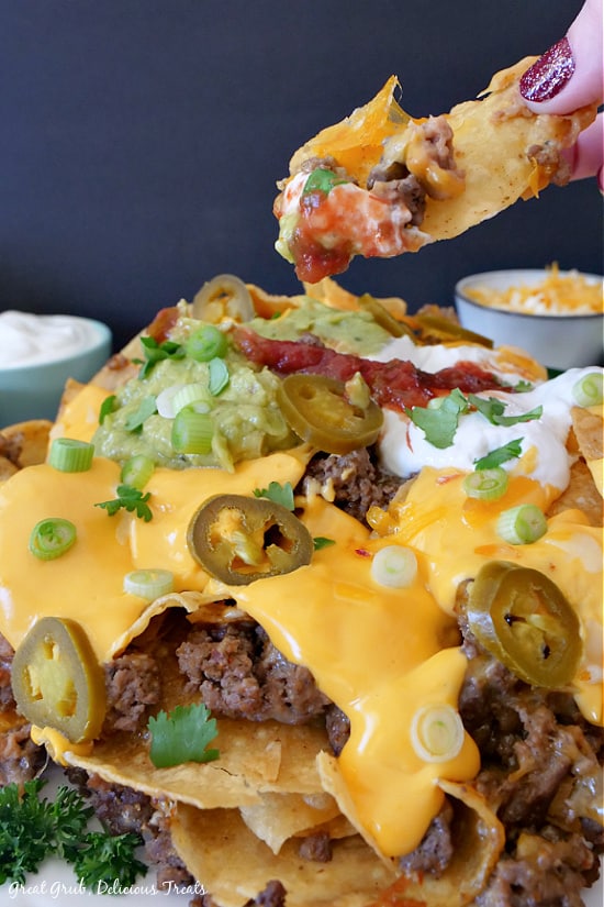 Mile High Nachos are a delicious appetizer recipe loaded with deliciously seasoned ground beef, refried beans, two types of shredded cheese and nacho sauce, and more. Mile High Nachos stacked on a place showcasing ground beef, nacho cheese, green onions, jalapenos, sour cream, salsa, and refried beans. Two small bowls with sour cream and shredded cheesed are in the background.