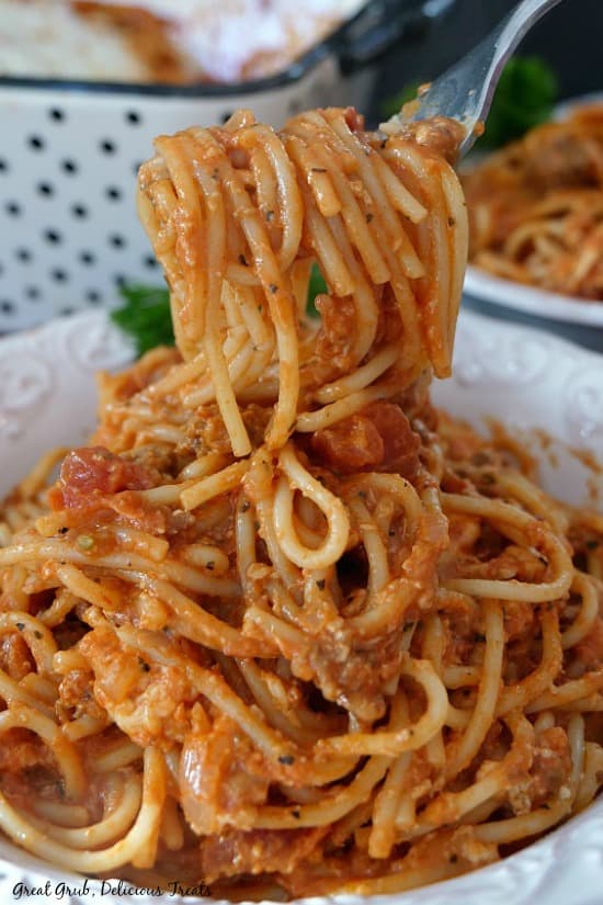 Italian Sausage Spaghetti Bake is a filling and delicious pasta recipe, loaded with Italian sausage, cheese, cream cheese and a homemade sauce. A fork with Italian Sausage Spaghetti Bake being held up over the white serving bowl filled with the spaghetti bake.