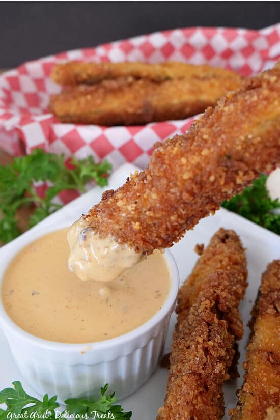 A close up photo of a zucchini strip being dipped in a sauce with more zucchini in the background.