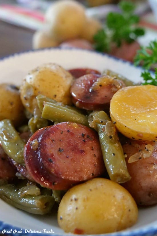 Sausage Green Bean Potato Casserole is seasoned perfectly, loaded with sausage kielbasa, green beans and baby potatoes. A close up photo of a serving of sausage, green beans and potatoes in a white bowl with blue trim.