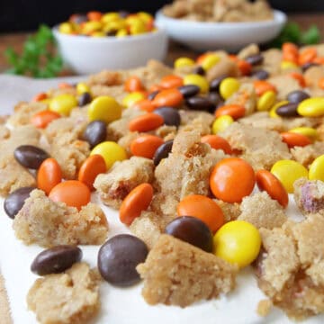 A white surface with a peanut butter cookie with a layer of cream cheese, and crumbled cookies and Reese's Pieces on top.