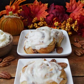 A wood surface with pumpkin cinnamon rolls on white square plates.