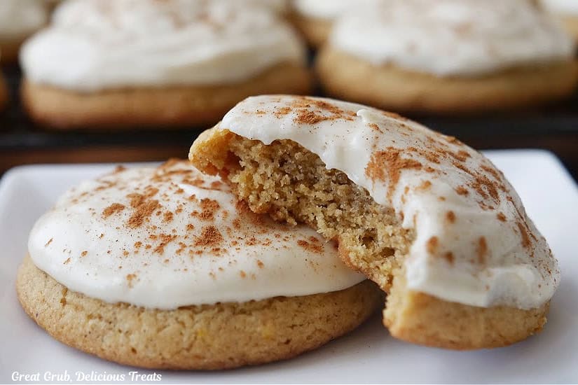 A white plate with two pumpkin cookies with brown sugar frosting on top with one bite taken out of one of the cookies.