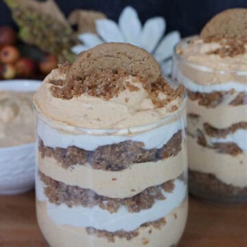 A wood surface with two glasses filled with pumpkin mousse, whipped cream, and gingersnap crumbs.