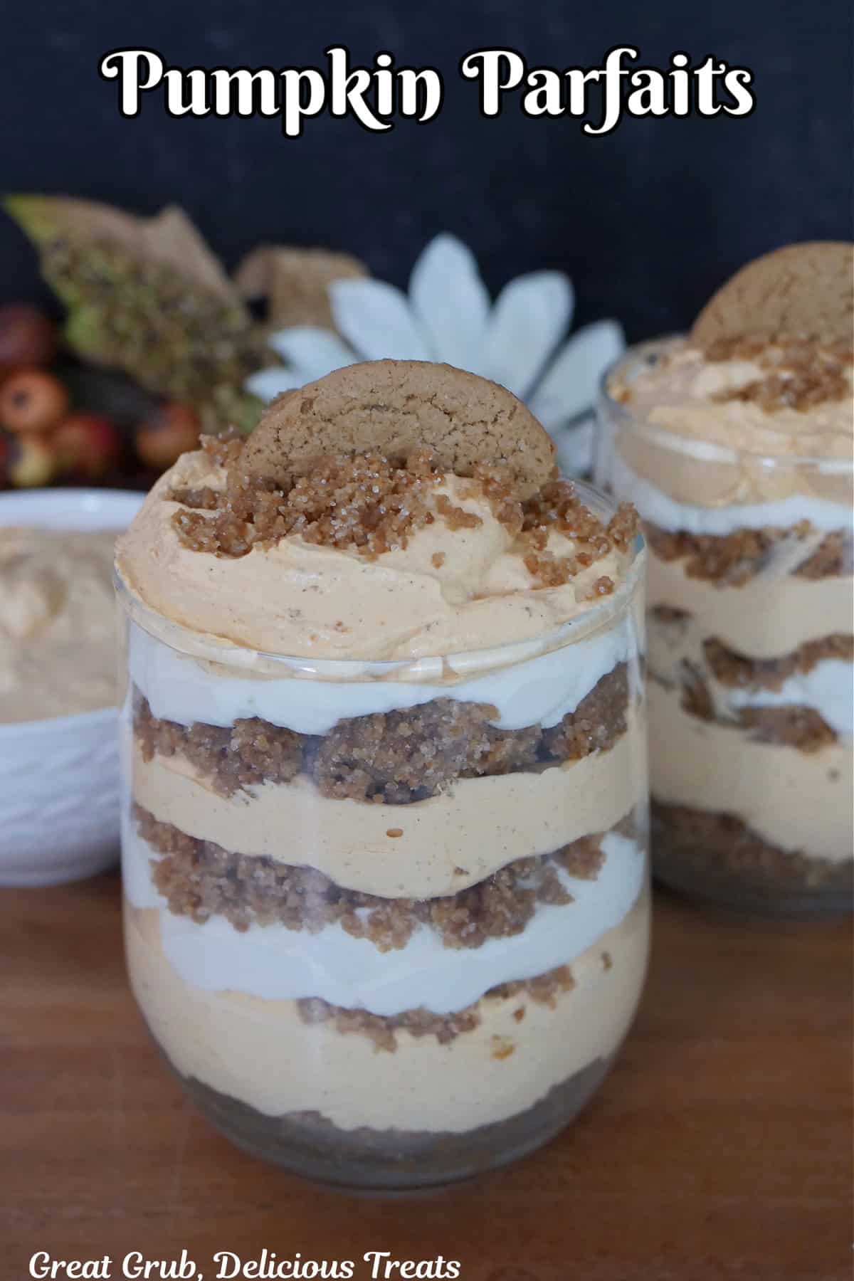 A wood surface with two glasses filled with pumpkin mousse, whipped cream, and gingersnap crumbs.
