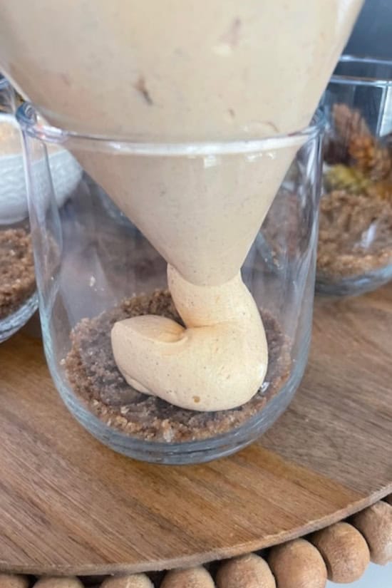 A wood surface with pumpkin mousse being added on top of gingersnap crumbs.