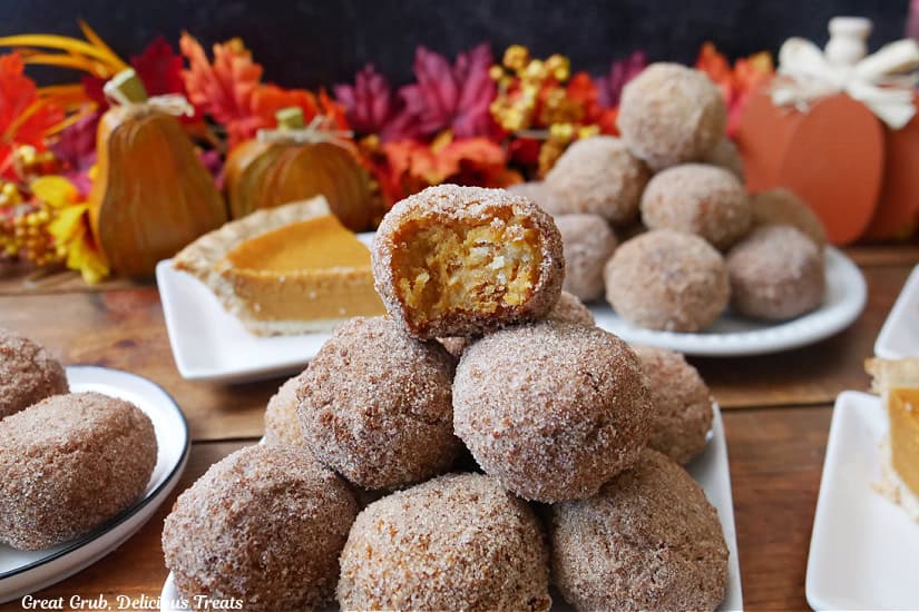 A wood surface with white plates with cinnamon sugar coated pumpkin pie balls stacked on each other, with fall foliage in the background