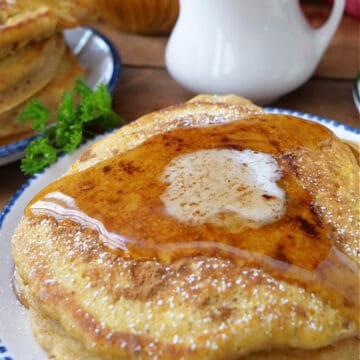A round white plate with blue trim with three fluffy pumpkin spice pancakes on it, topped with a pad of butter, and syrup.