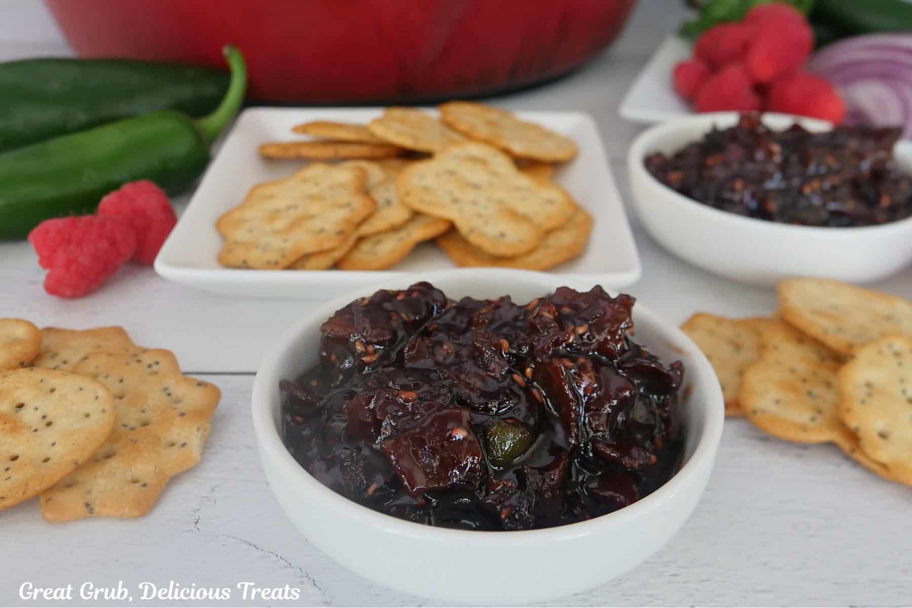 A white surface with two white bowls filled with raspberry jalapeno bacon jam with crackers placed around the bowls.