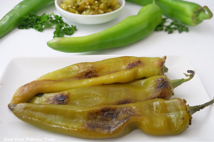 A white plate with three roasted green chiles on it, and three more green chiles in the background that have not been roasted yet.