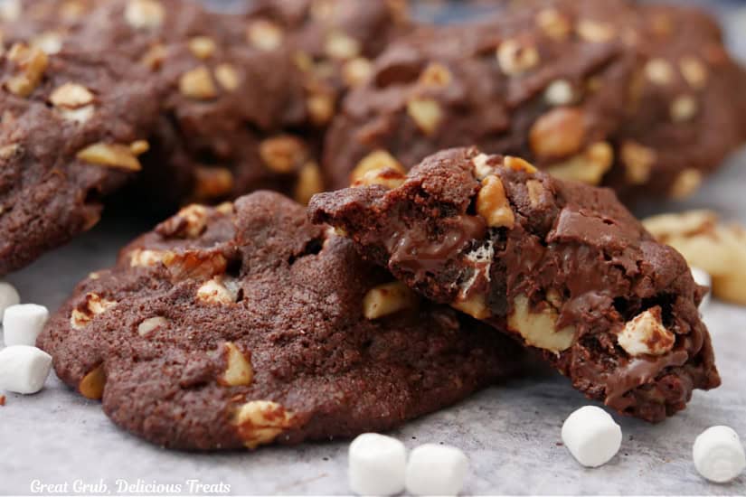 A white surface with rocky road dark chocolate cookies on it with a bite taken out of one of the cookies.