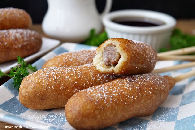 A horizontal photo of a light blue and white checkered square plate with four sausage pancake on a stick with a bite taken out of one of them.