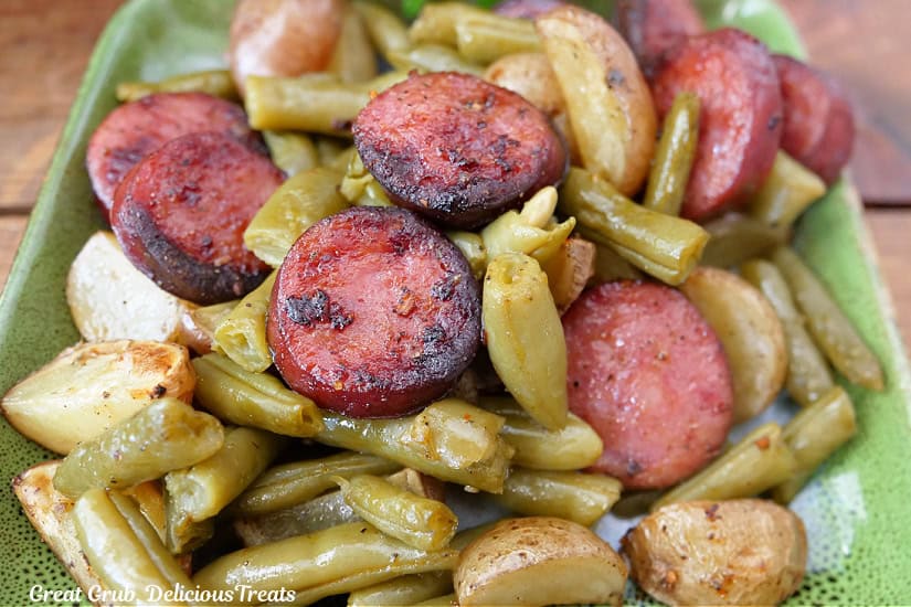 A square green plate with sliced smoked sausage, potatoes, and green beans on it.