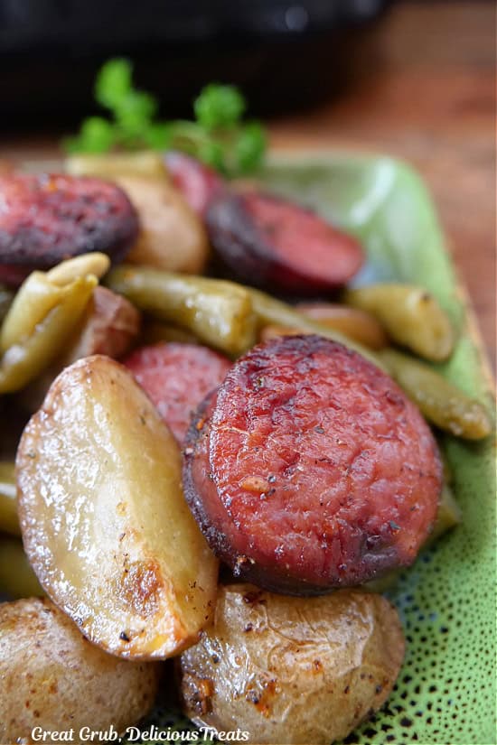 A close up photo of a green plate with potatoes, green beans and sliced smoked sausage on it.
