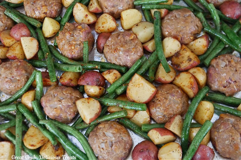 A foil lined baking sheet with meatballs, potatoes, and green beans on it before being baked.