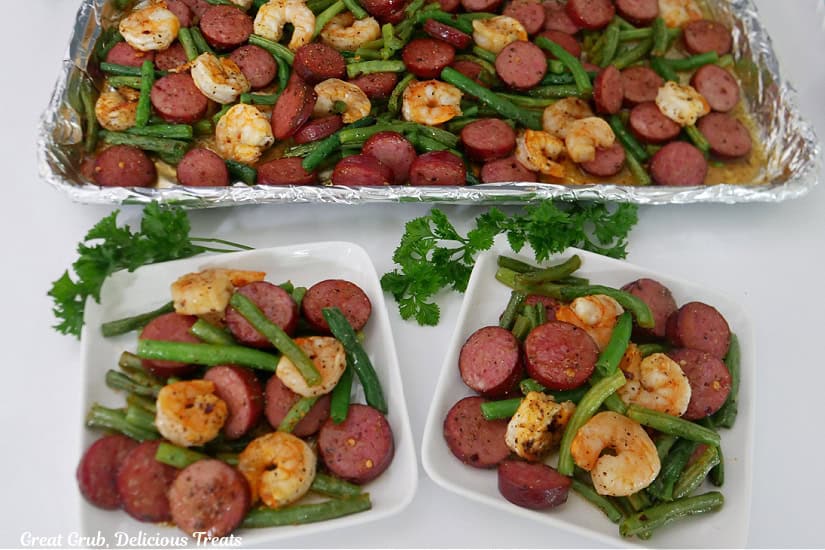 A white surface with two square plates and a sheet pan filled with sausage, green beans and shrimp.