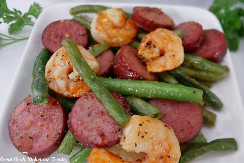 A white square plate with a serving of sausage green beans and shrimp.