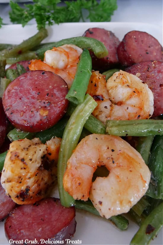 A close up photo of sliced smoked sausage, fresh green beans, and shrimp on a white plate.