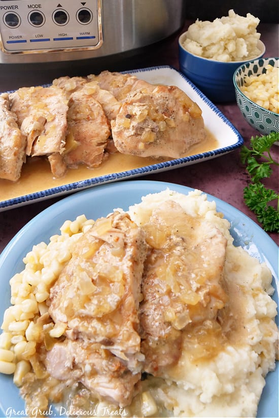 Slow Cooker Pork Chops with Apples are an easy pork chop recipe that is easily made right in the crock pot. A plate with potatoes, corn, and pork chops, with a long white plate lined with pork chops in the background.