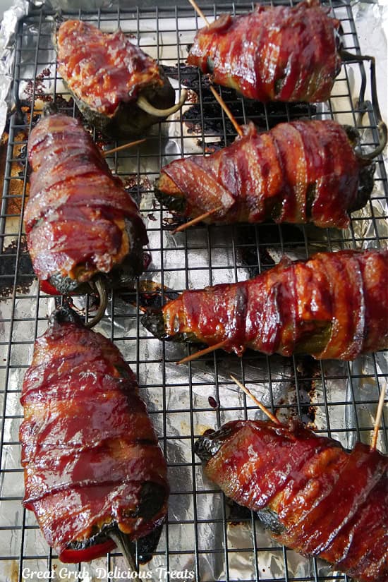 A wire rack on a foil lined baking sheet with seven bacon wrapped smoked stuffed poblano peppers with toothpicks still sticking out of them after being smoked.