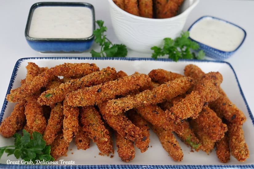 A white rectangle plate with blue trim with spicy green beans on it with two blue square bowls filled with ranch dipping sauce.