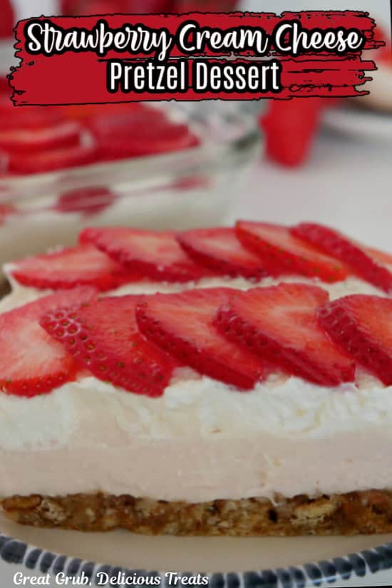 A white plate with blue trim with a slice of strawberry pretzel dessert on it with fresh strawberries in the background.