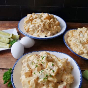 A wood surface with three white bowls filled with potato salad, with a small white plate with pickle spears, and two hard boiled eggs, and the title of the recipe in text at the top.