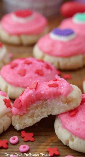 A close up of mini sugar cookies with a bite taken out of one of them. They have pink buttercream frosting and candy sprinkles on top.