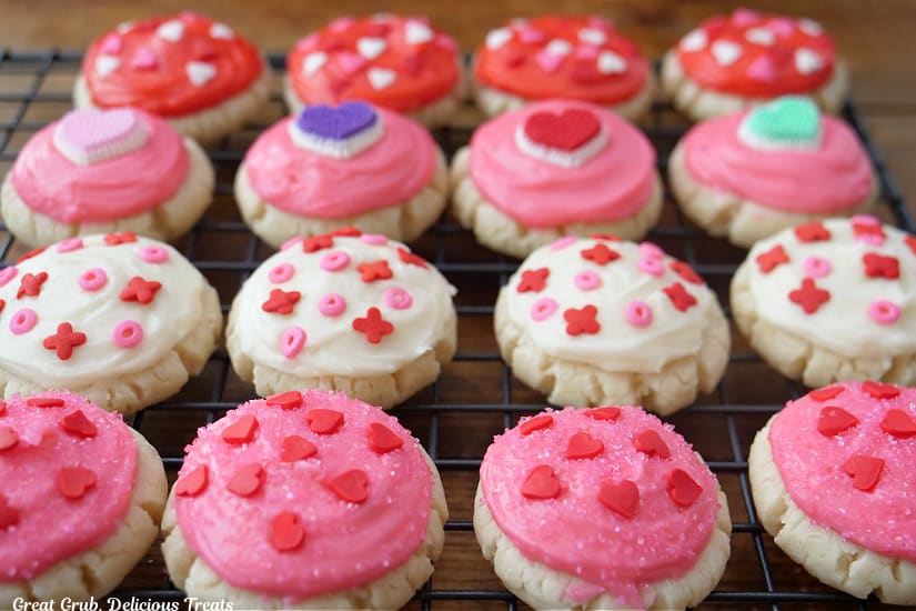 A wire rack with sixteen mini sugar cookies with pink, white, and red buttercream frosting and candy sprinkles on them.