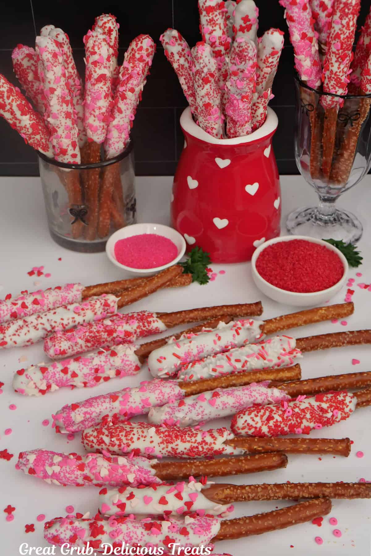 A white surface with white chocolate caramel coated pretzel rods placed on it and more in put in decorated cups.