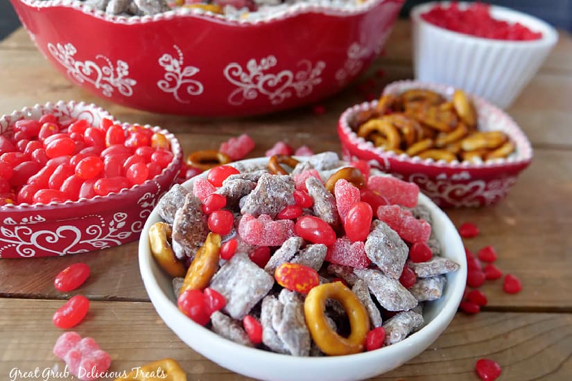 Valentine Puppy Chow Mix is a simple recipe to make that is perfect for cinnamon lovers. A white bowl with valentine muddy buddy mix in it with two red heart bowls filled with pretzels and red hot candies.